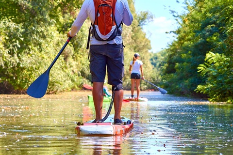 licensed stock photo of a man standing on a paddleboard on a river with a woman on a paddleboard ahead in the distance