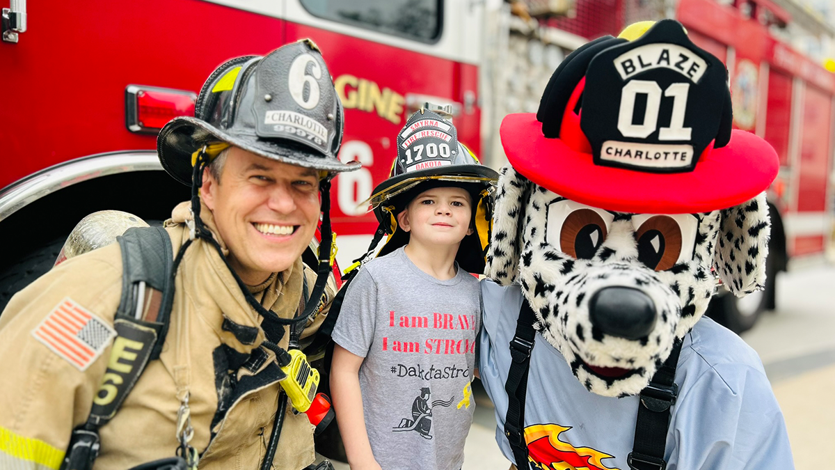 Dakota stands with Firefighter William Roosenberg and Charlotte Fire's mascot, Blaze, enjoying the celebration in front of Engine 6.