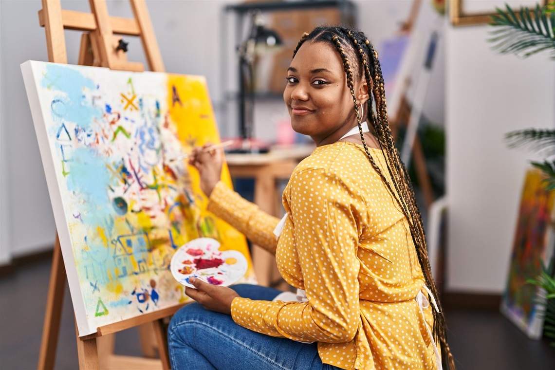 A young woman looks back at the camera while painting her next masterpiece on the easel in front of her.