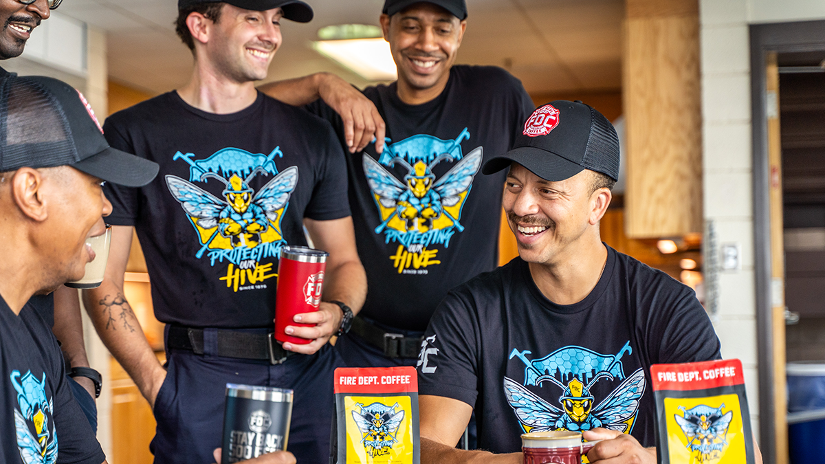 Five Charlotte firefighters gather at a table with bags of their Fire Dept. Coffee. They are all smiling and wearing shirts with the coffee's logo.