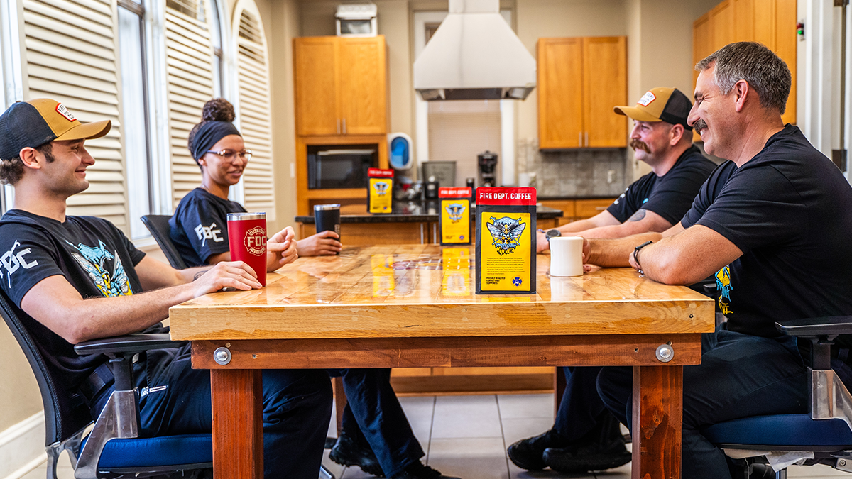 Four Charlotte firefighters sit together at a table and smile at each other. There are several bags of their coffee around, and all of the firefighters have a cup of their own.