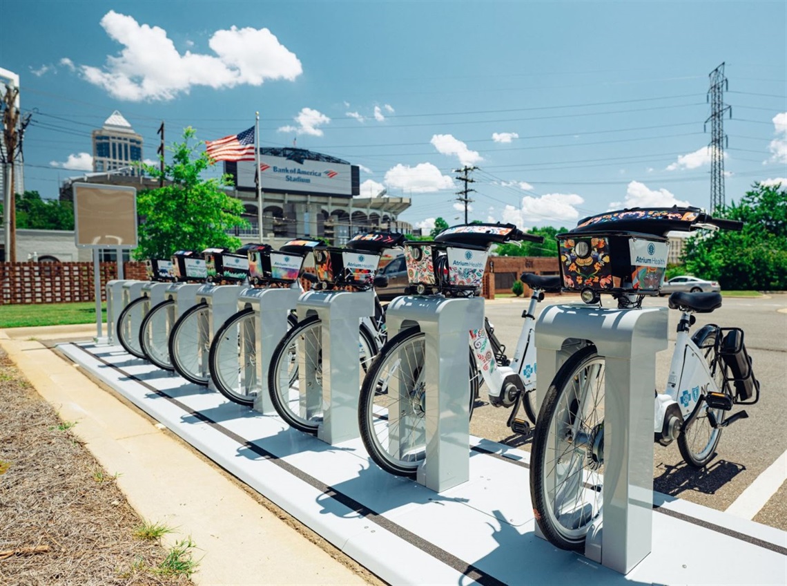 Joy Rides bikes lined up in a rack with Bank of America Stadium behind them. It's a bright, sunny day with a smattering of clouds.