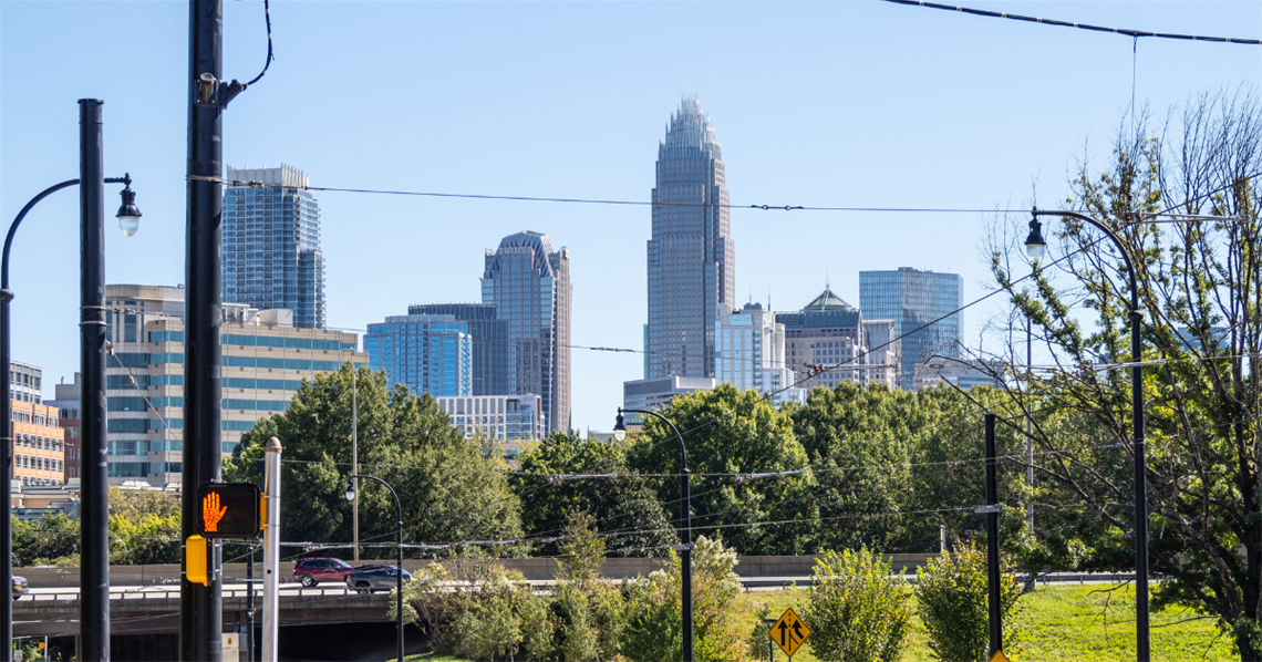 Charlotte skyline viewed from the Beatties Ford Corridor of Opportunity. Green trees and cars traveling over a bridge are in the foreground.