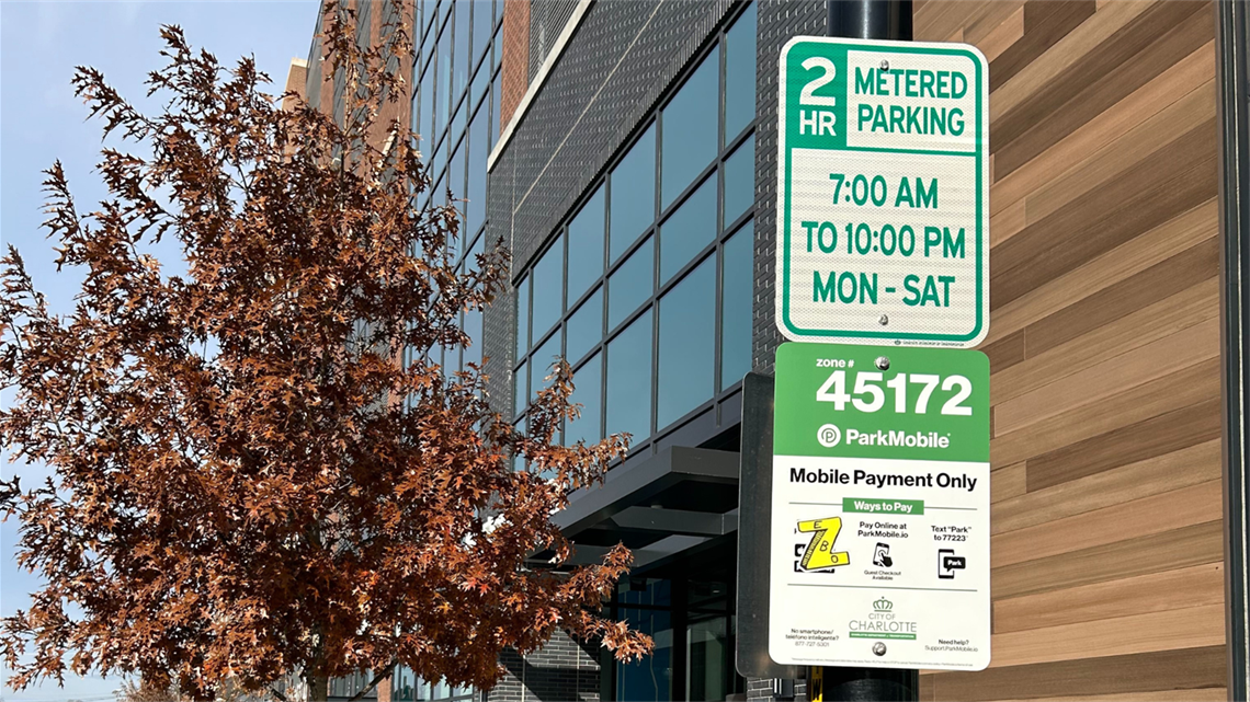 Parking sign in Uptown Charlotte with hours and instructions for payment clearly stated. A tree with browning leaves and a modern building appears behind the signage.