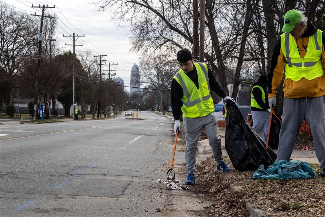 Keep Charlotte Beautiful volunteers clean up litter on a Charlotte street. The skyline is visible behind them.