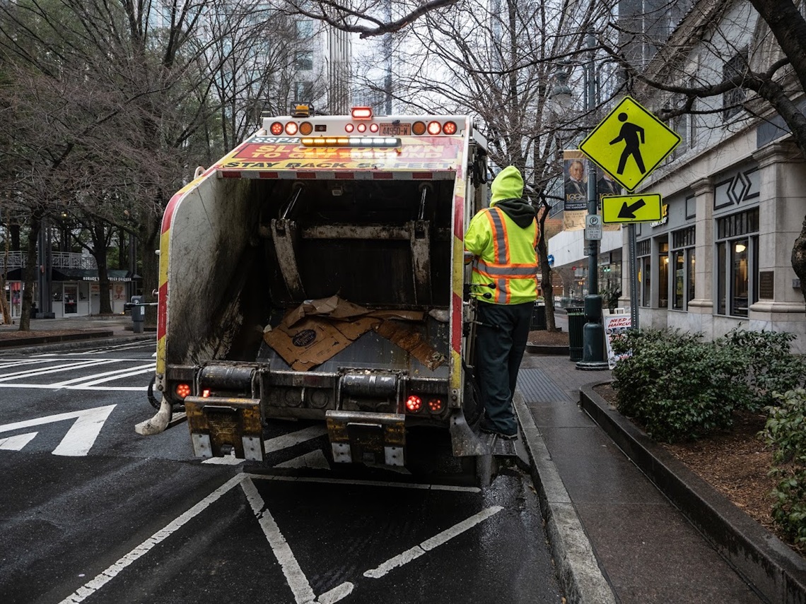 A Solid Waste Services employee rides on the back of a truck during a collection route in Uptown.