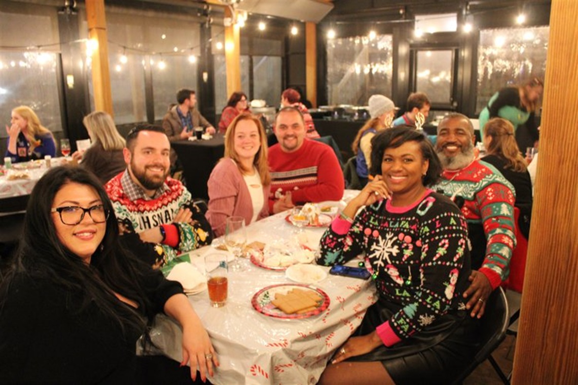 A diverse group of people gather around a table in Christmas sweaters.