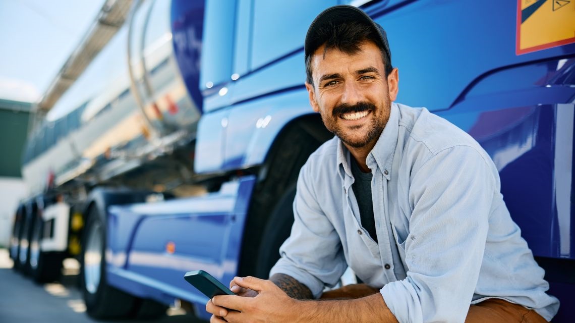 A man smiles with his phone in hand outside of his parked commercial truck.